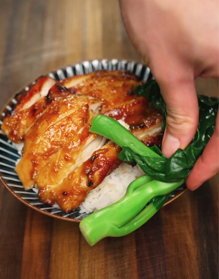 A hand places freshly steamed bok choy into a bowl of miso-glazed chicken over rice, completing the colorful and balanced meal presentation.
