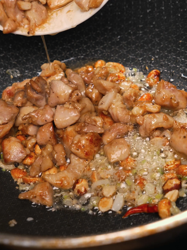 Marinated chicken is being added to a hot skillet filled with chopped garlic, chilies, and ginger, sizzling in oil to build the flavor base for Kung Pao Chicken. The aromatics are beginning to brown and release their aroma.