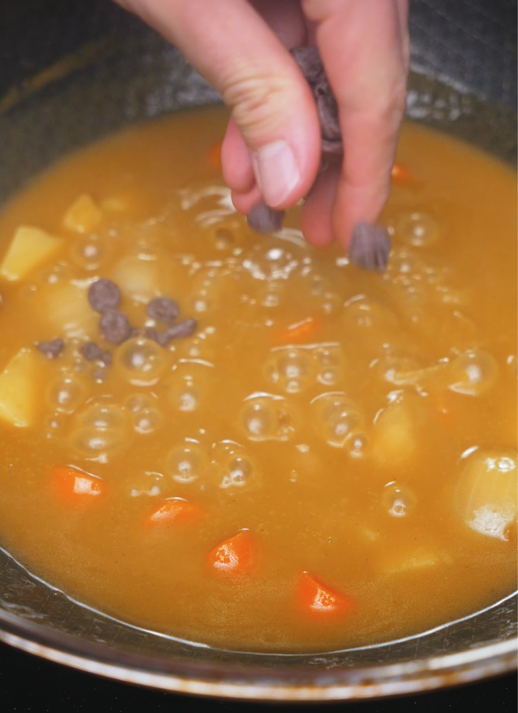 Hand adding dark chocolate chips to bubbling Japanese curry sauce with potatoes and carrots – rich and savory chicken katsu curry base.