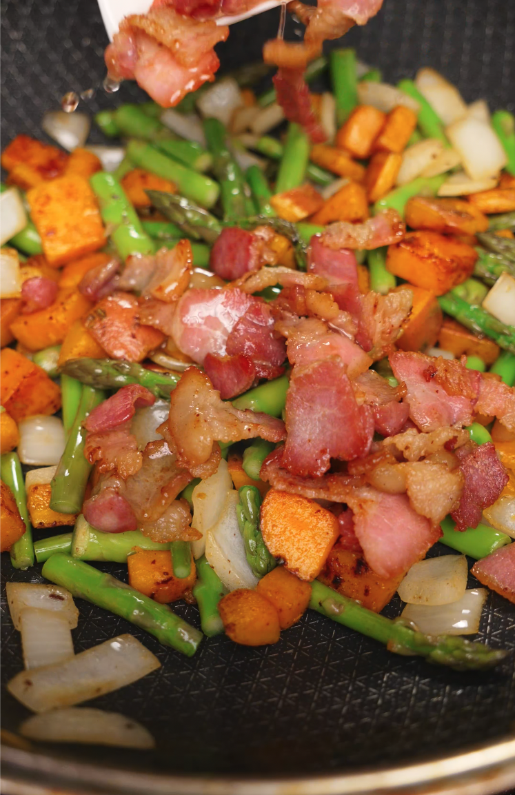Crisped bacon pieces are added to a colorful stir-fry of sweet potatoes, asparagus, and onions in a hot pan. The combination of textures and savory aromas builds the perfect base for a hearty steak side dish.