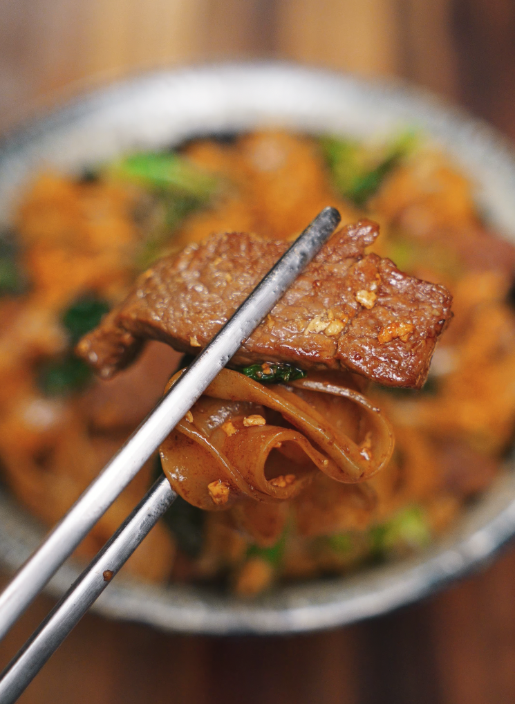 A close-up of chopsticks holding a piece of caramelized beef and twisted saucy rice noodles from Pad See Ew. The glossy texture highlights the perfect stir-fry sear and balance of flavors.