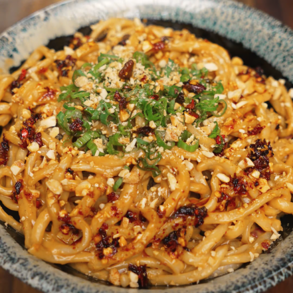 Final dish shot of spicy peanut noodles topped with chili oil, scallions, and crushed peanuts in a speckled ceramic bowl. The vibrant topping adds crunch and heat, making this bowl pop with texture and flavor.