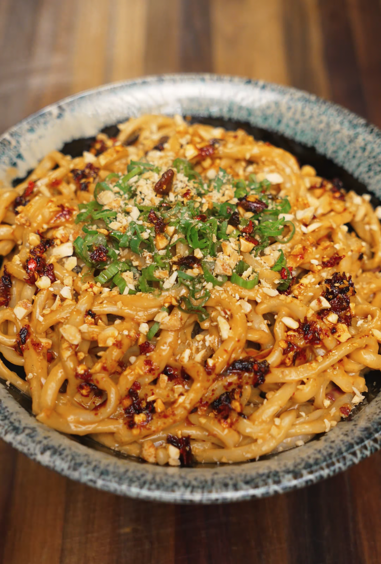 Final dish shot of spicy peanut noodles topped with chili oil, scallions, and crushed peanuts in a speckled ceramic bowl. The vibrant topping adds crunch and heat, making this bowl pop with texture and flavor.