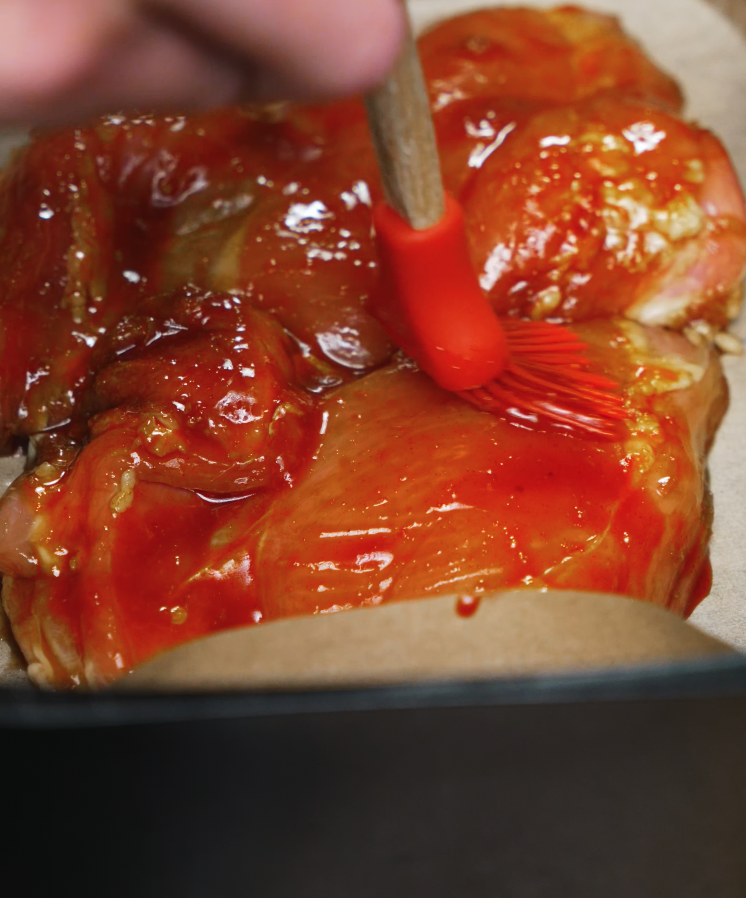 Brushing a vibrant gochujang barbecue marinade onto raw chicken thighs in a baking tray lined with parchment paper. The rich red glaze is thick and glossy, coating the chicken evenly to prepare it for roasting or air frying. This step ensures a flavorful, sticky, and caramelized crust in the final BBQ gochujang chicken dish—perfect for meal prep or weeknight dinners.