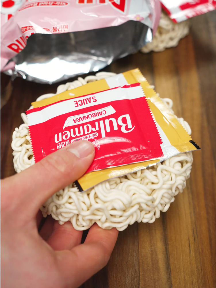 A hand holding a brick of uncooked instant ramen noodles with a Buldak Carbonara sauce packet on top, highlighting the convenience and brand used for this spicy, cheesy Korean-style instant carbonara ramen recipe.