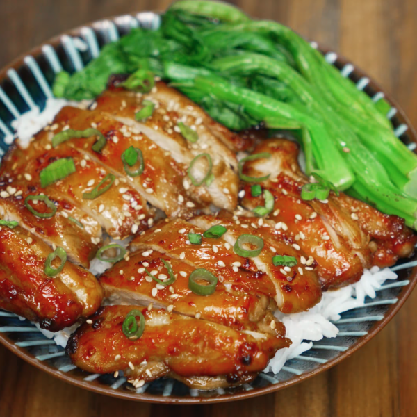 Bowl of sliced chicken bulgogi served over steamed rice, topped with sesame seeds and scallions, alongside sautéed greens.