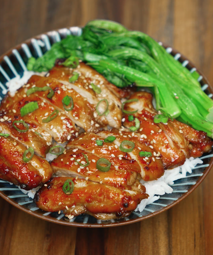 Bowl of sliced chicken bulgogi served over steamed rice, topped with sesame seeds and scallions, alongside sautéed greens.