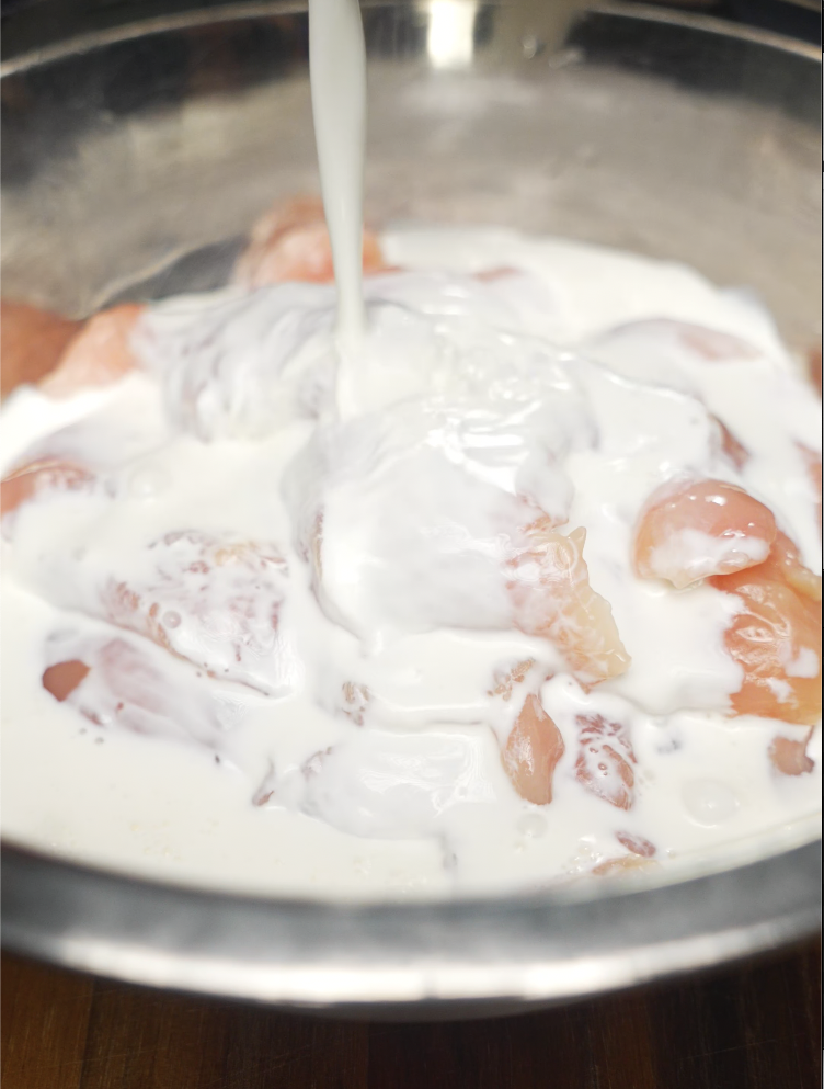 Raw chicken tenders soaking in a bowl of creamy buttermilk, prepping for deep frying. The marinade tenderizes the meat and helps the flour coating stick, ensuring an ultra-crispy bite before being coated in honey glaze—essential for fried chicken lovers or Southern recipe roundups.