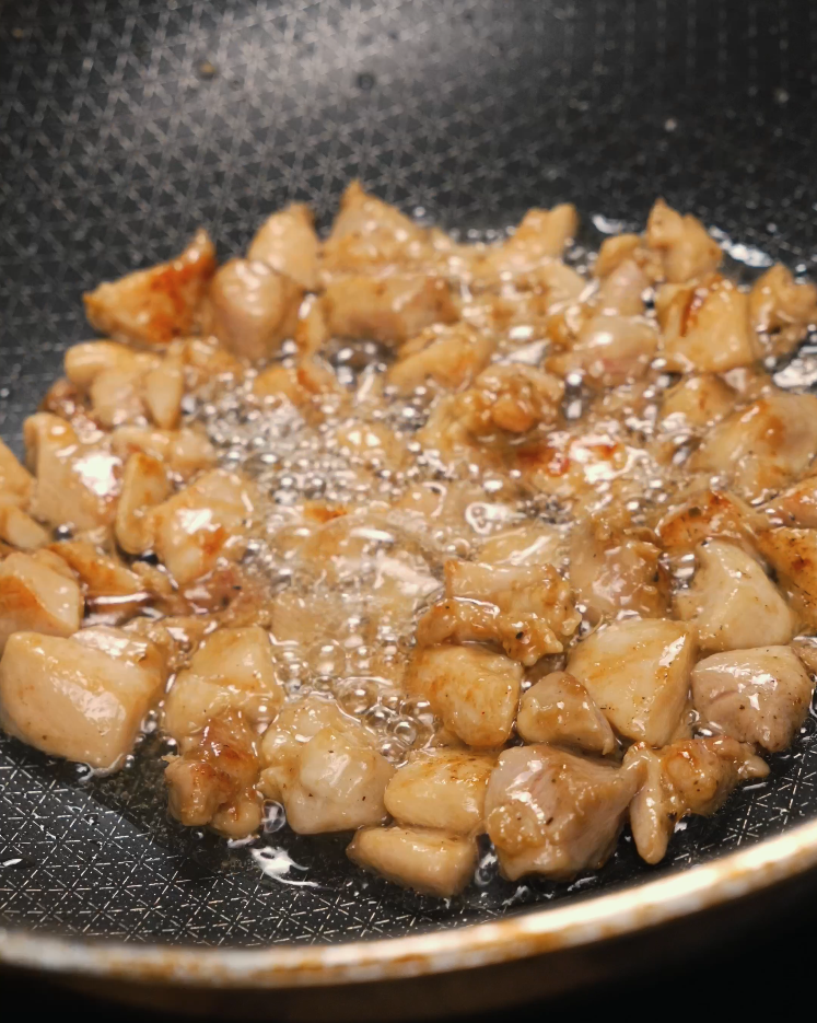Bite-sized pieces of chicken thigh searing in bubbling oil inside a frying pan, developing a golden crust. The chicken is being browned before the sauce and peanuts are added, building layers of flavor.