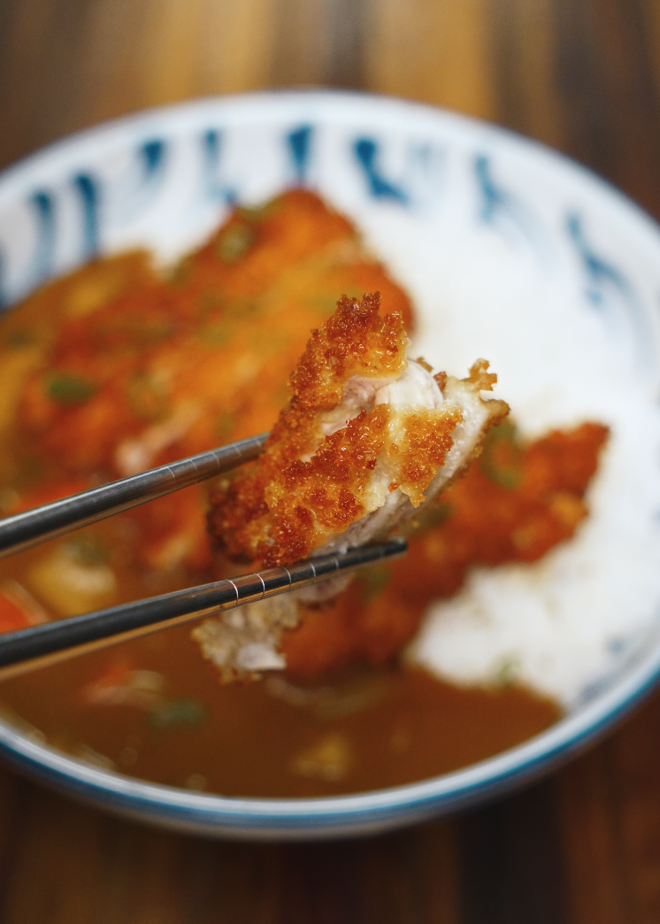 Close-up of crispy chicken katsu held by chopsticks above a bowl of curry and rice – crunchy and tender bite of chicken katsu curry.