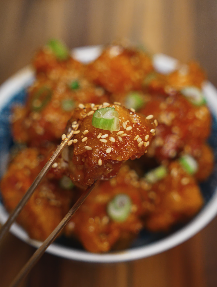 A close-up of crispy Korean fried chicken coated in a sticky soy garlic glaze, topped with toasted sesame seeds and fresh scallions, held between metal chopsticks with the rest of the bowl blurred in the background.