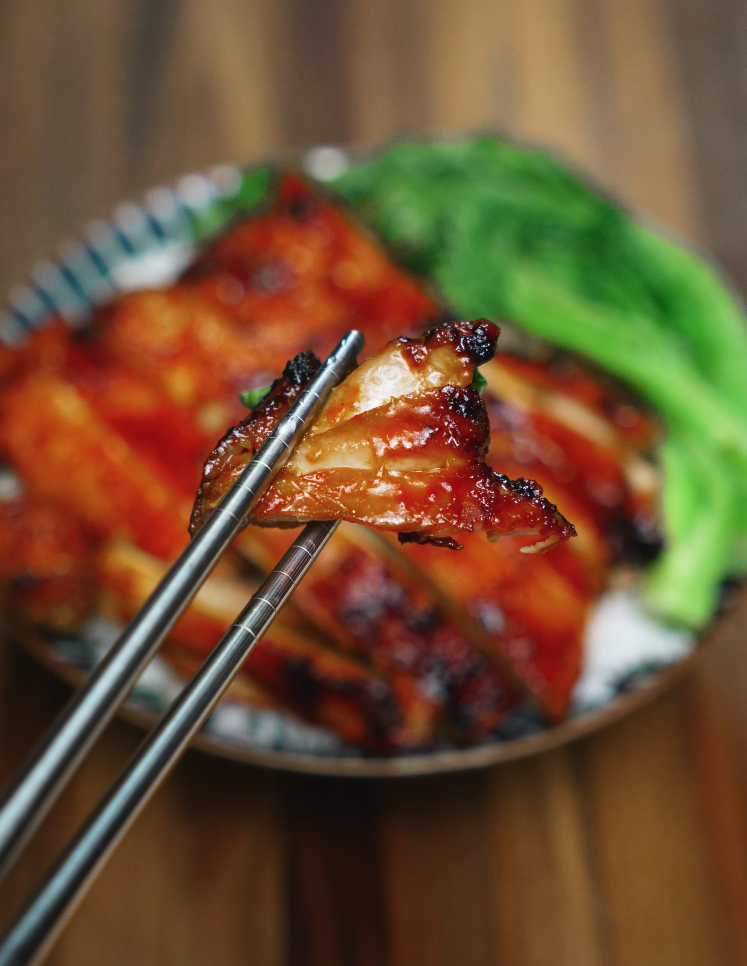 A glossy, perfectly caramelized piece of BBQ gochujang chicken lifted by metal chopsticks over a full rice bowl. The charred edges highlight the umami-rich glaze, while the tender meat shows off juicy, flavorful layers. This close-up shot showcases the irresistible texture and bold Korean-inspired flavors that define this gochujang chicken recipe.