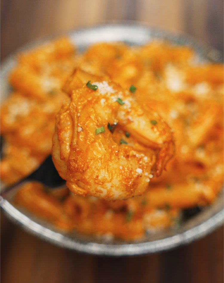 A plump shrimp coated in creamy, spicy sauce is held by a fork above a bowl of rigatoni. The shrimp is tender, golden, and dusted with parmesan and herbs for a flavorful finish.