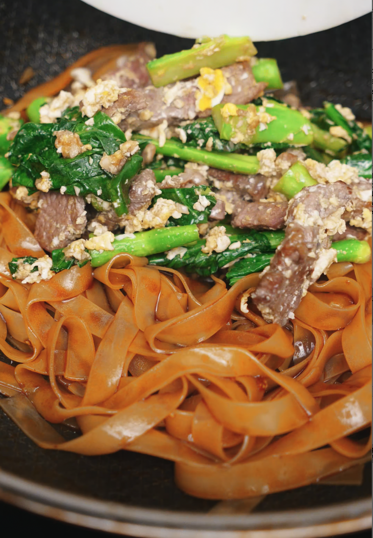 Tender stir-fried beef and Chinese broccoli (gai lan) being added to a hot wok with wide rice noodles coated in a rich soy-based sauce. This step brings all Pad See Ew components together.