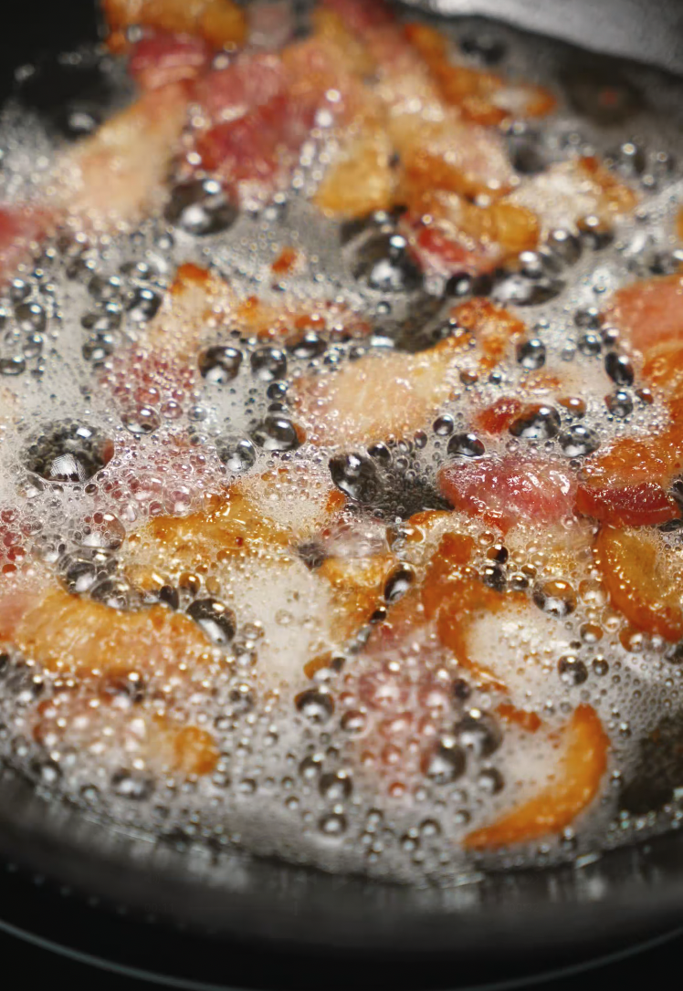 Thick slices of bacon sizzling in a hot pan, bubbling in their own rendered fat to achieve a crispy, golden-brown finish that will later be used to enhance the flavor of the instant carbonara ramen recipe.