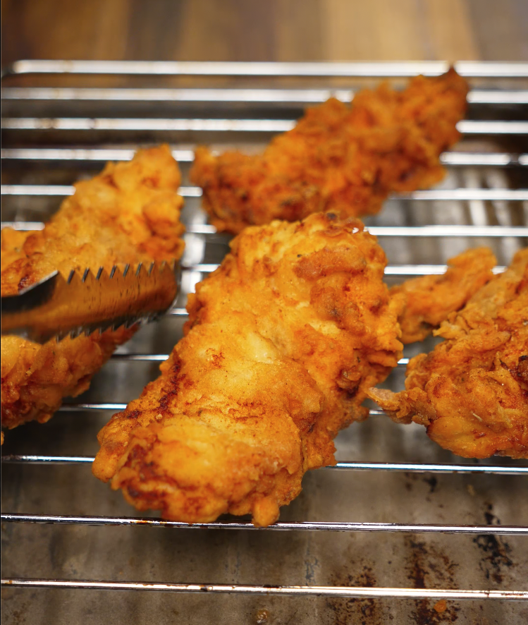 Freshly fried honey butter chicken tenders cooling on a wire rack, showcasing their ultra-crispy, golden-brown breading. These tenders are the perfect base for a sticky honey butter glaze, making them ideal for comfort food recipes, chicken tender tutorials, or crispy fried chicken content.