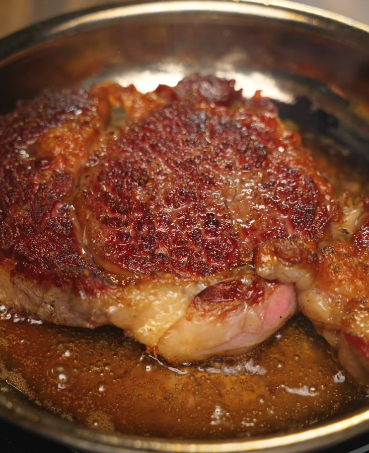 Close-up of a ribeye steak from Asian Style Steak Frites with a caramelized crust sizzling in a pan with melted butter.