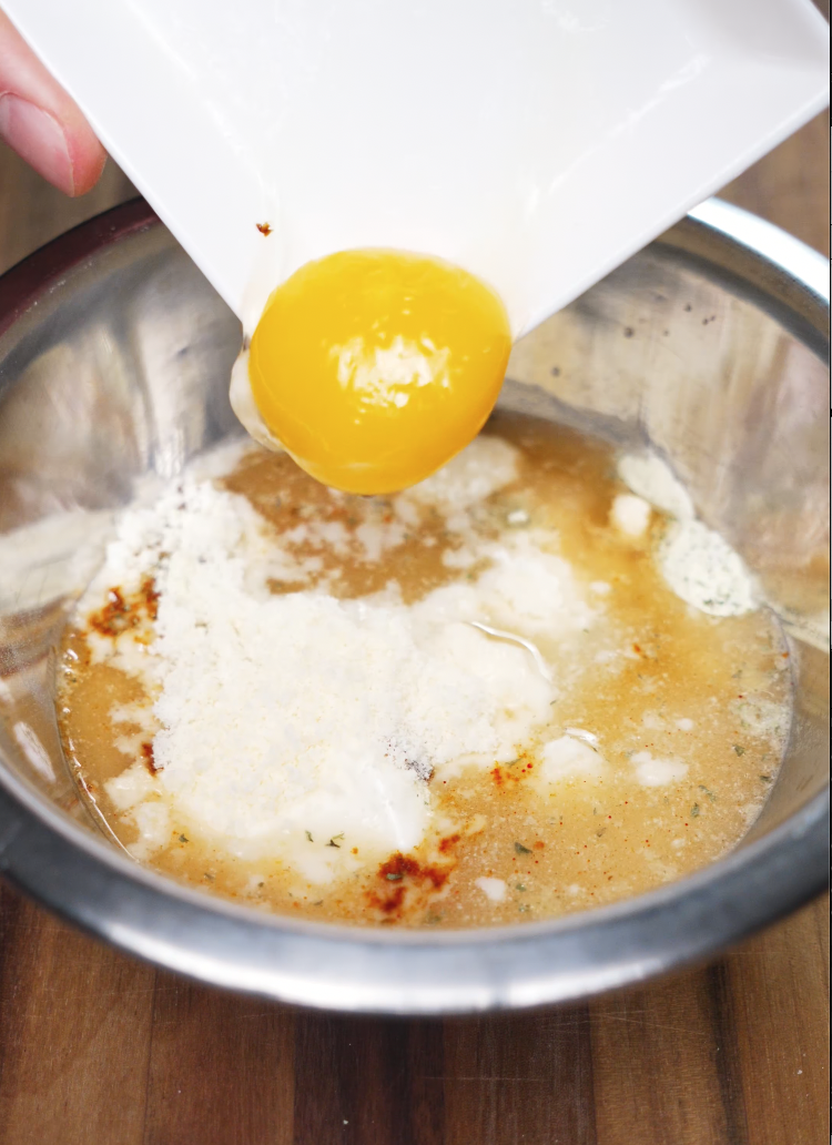 A raw egg yolk sliding into a bowl filled with grated cheese, pasta water, and seasonings, forming the rich, creamy base for a homemade instant carbonara ramen sauce, adding silky texture and umami depth.