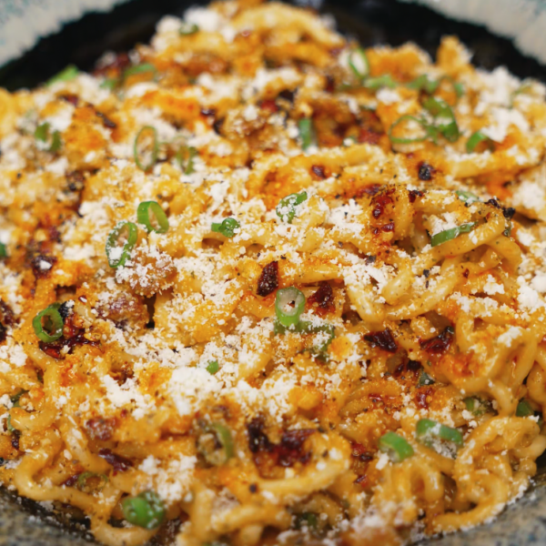 A large bowl of instant carbonara ramen topped generously with parmesan cheese and chopped scallions, featuring crispy bacon and slightly charred edges for an ultra-comforting and flavorful instant noodle upgrade.