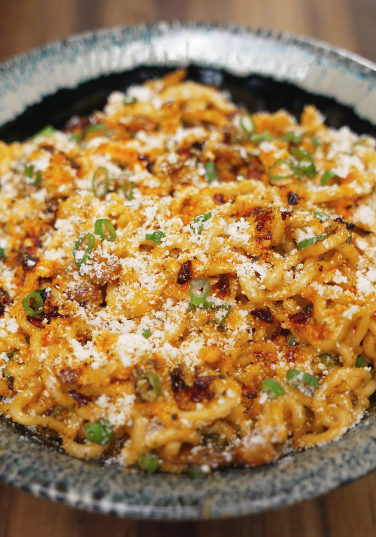 A large bowl of instant carbonara ramen topped generously with parmesan cheese and chopped scallions, featuring crispy bacon and slightly charred edges for an ultra-comforting and flavorful instant noodle upgrade.