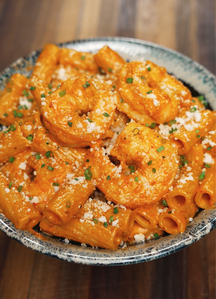 A bowl of rigatoni tossed in a spicy, creamy sauce with perfectly cooked shrimp, finished with parmesan and chives. The vibrant orange color and creamy texture make this a bold, comforting pasta dish.