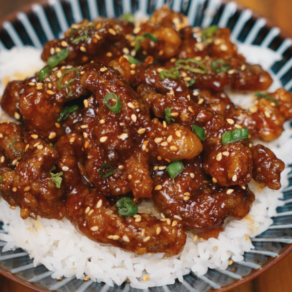 A full serving of sticky crispy beef piled on top of white rice in a patterned bowl, garnished with sesame seeds and chopped green onions for contrast and crunch.