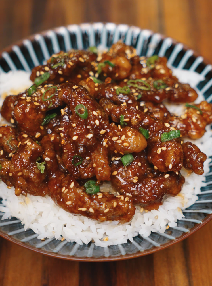 A full serving of sticky crispy beef piled on top of white rice in a patterned bowl, garnished with sesame seeds and chopped green onions for contrast and crunch.
