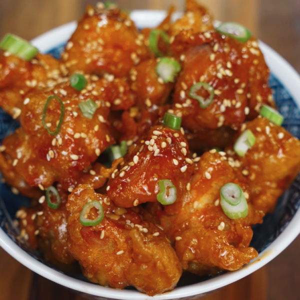 A bowl of perfectly fried and glazed Korean chicken bites tossed in soy garlic sauce, generously sprinkled with sesame seeds and sliced scallions, served in a blue and white ceramic bowl on a wooden table.