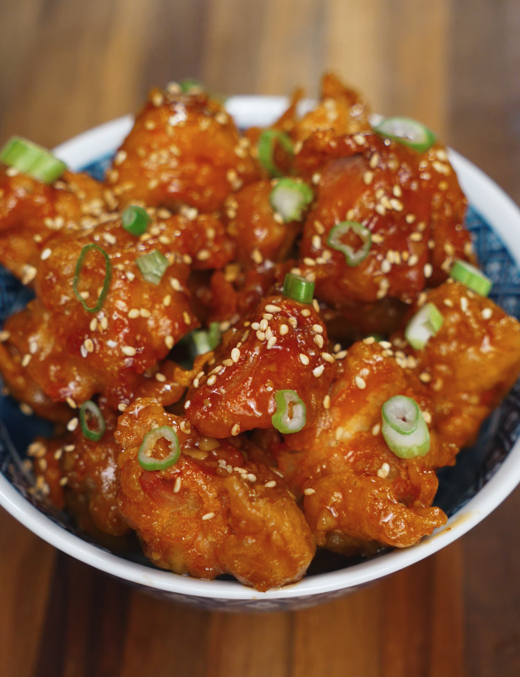 A bowl of perfectly fried and glazed Korean chicken bites tossed in soy garlic sauce, generously sprinkled with sesame seeds and sliced scallions, served in a blue and white ceramic bowl on a wooden table.
