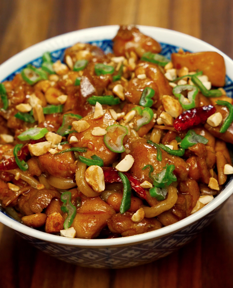 A full bowl of Kung Pao Chicken Noodles, topped with crushed peanuts, scallions, and dried chilies. The noodles are coated in a sticky, savory sauce with caramelized chicken pieces and crunchy peanuts throughout.
