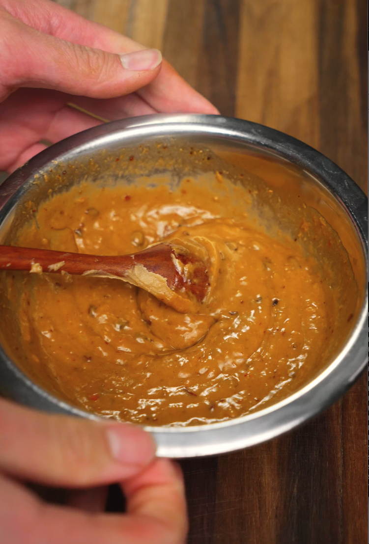 A wooden spoon stirs together a thick, spicy peanut sauce with visible chili flakes and crushed aromatics in a metal bowl. The sauce is glossy and smooth, ready to coat freshly cooked noodles with bold, nutty heat.