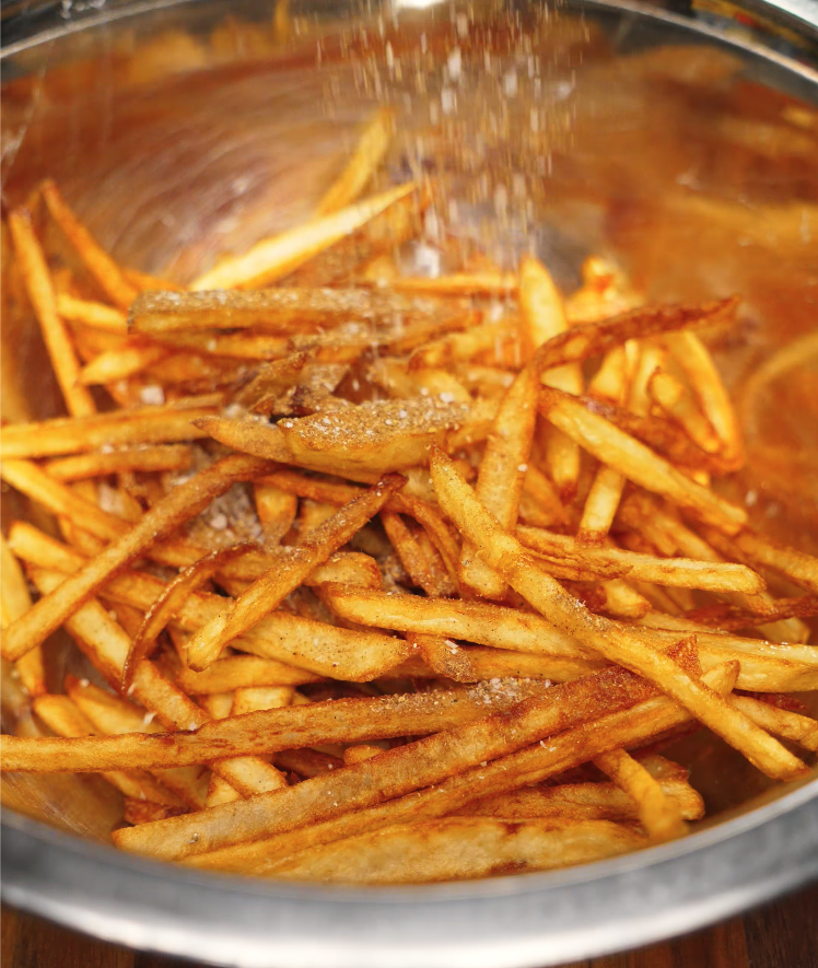 Freshly fried thin-cut five-spice French fries being tossed and salted in a large metal mixing bowl.
