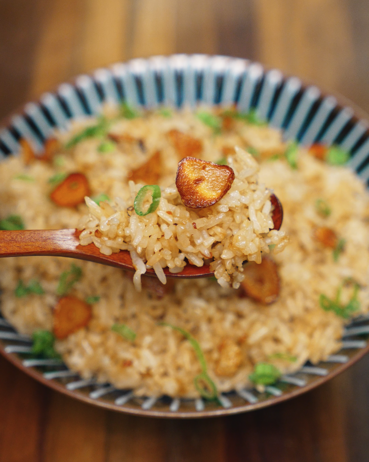 A close-up view of a wooden spoon lifting a perfect bite of garlic butter fried rice, featuring golden crispy garlic, fragrant rice, and vibrant green onions. The spoonful highlights the rich flavor and textural contrast of the dish, showcasing the ideal portion for a food blog or Asian dinner recipe post.