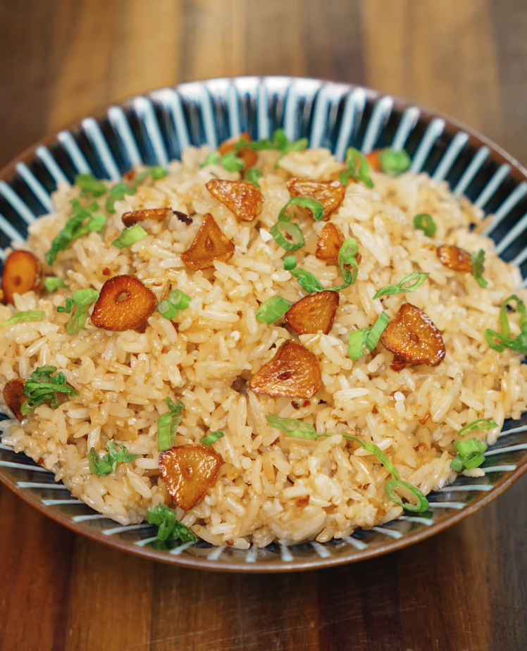 A finished bowl of garlic butter fried rice garnished with crispy golden garlic chips and chopped green onions. The fluffy, flavorful rice is glistening with garlic butter, delivering a savory and comforting dish ideal for meal prep or a quick weeknight dinner. This visually appealing bowl highlights the simplicity and bold flavors of a popular Asian comfort food.