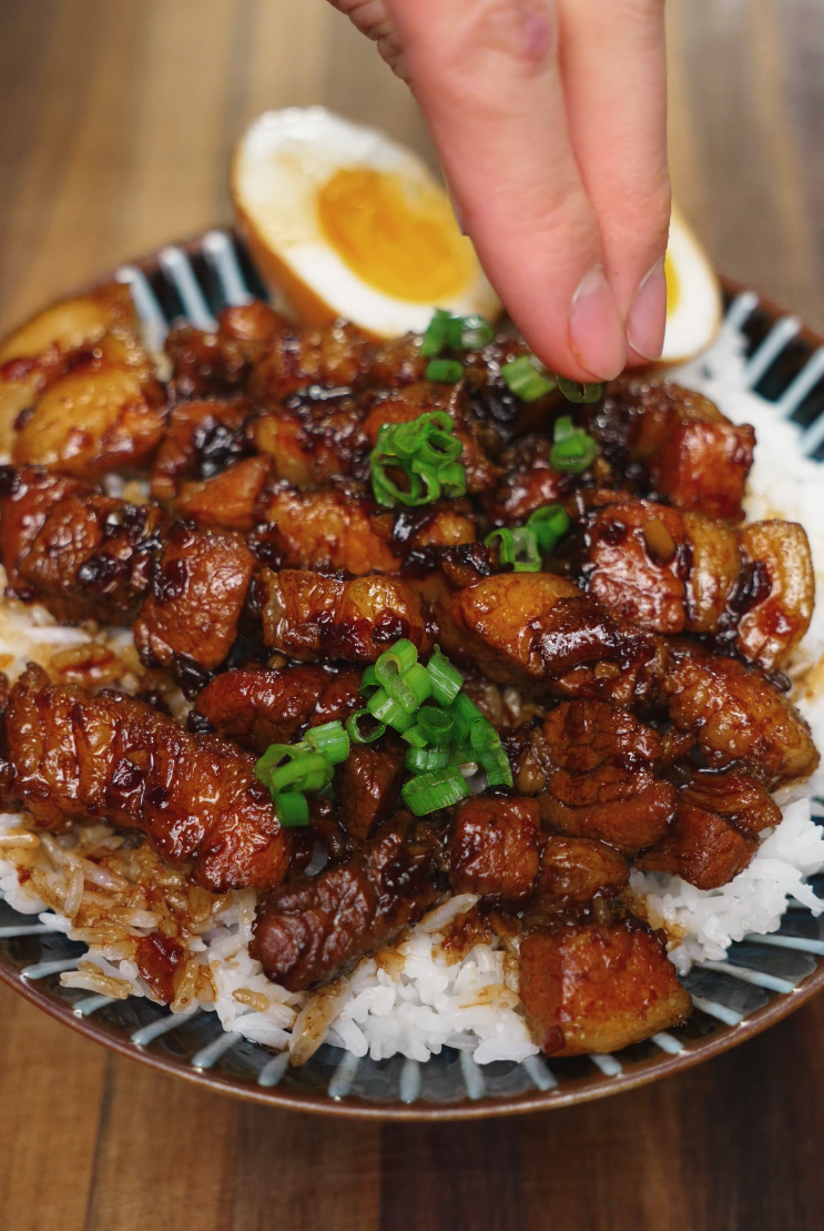 A close-up shot of a hand sprinkling fresh chopped green onions over a bowl of soy braised pork belly, served atop fluffy white rice and paired with a jammy soft-boiled egg for the perfect finishing touch.