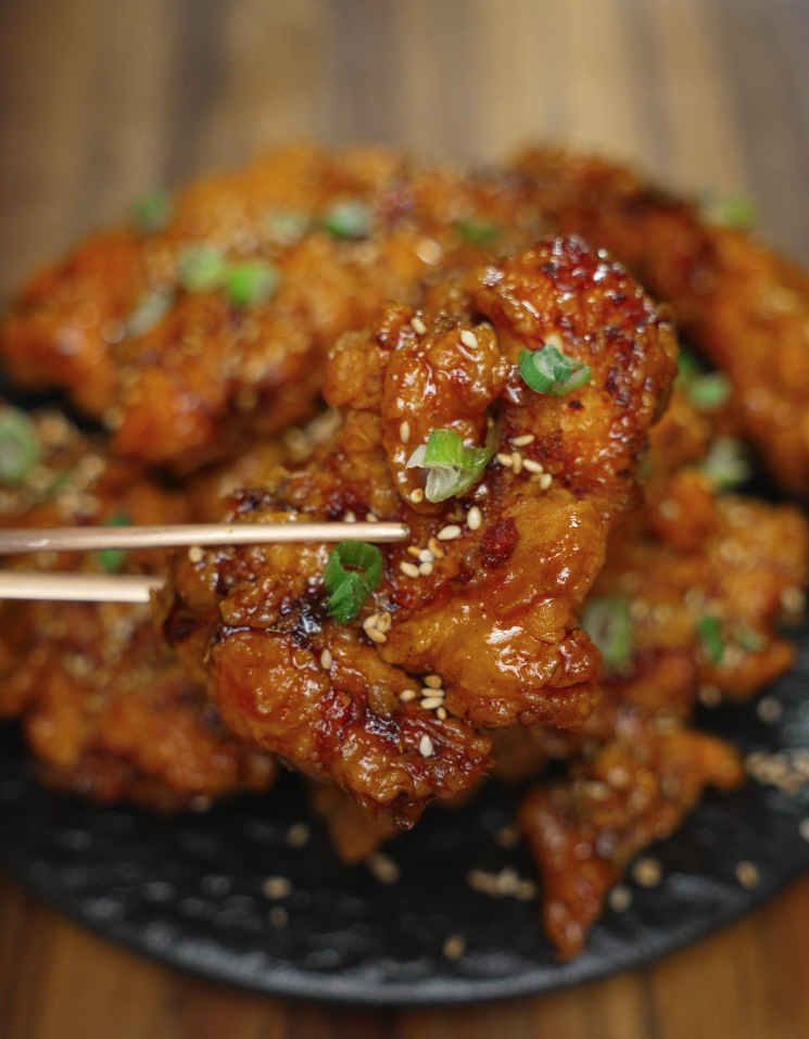 Close-up of a sticky, glazed honey butter chicken tender held with chopsticks, sprinkled with sesame seeds and garnished with sliced scallions. The crispy exterior peeks through the shiny glaze, making this bite look both crunchy and flavorful—perfect for promoting viral chicken recipes or Asian fusion comfort food.