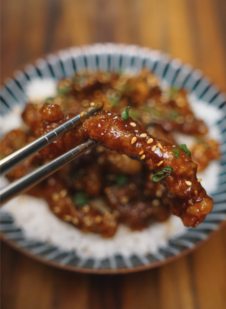 Chopsticks holding up a glistening piece of sticky crispy beef, coated in a thick soy garlic glaze and sprinkled with sesame seeds and scallions.