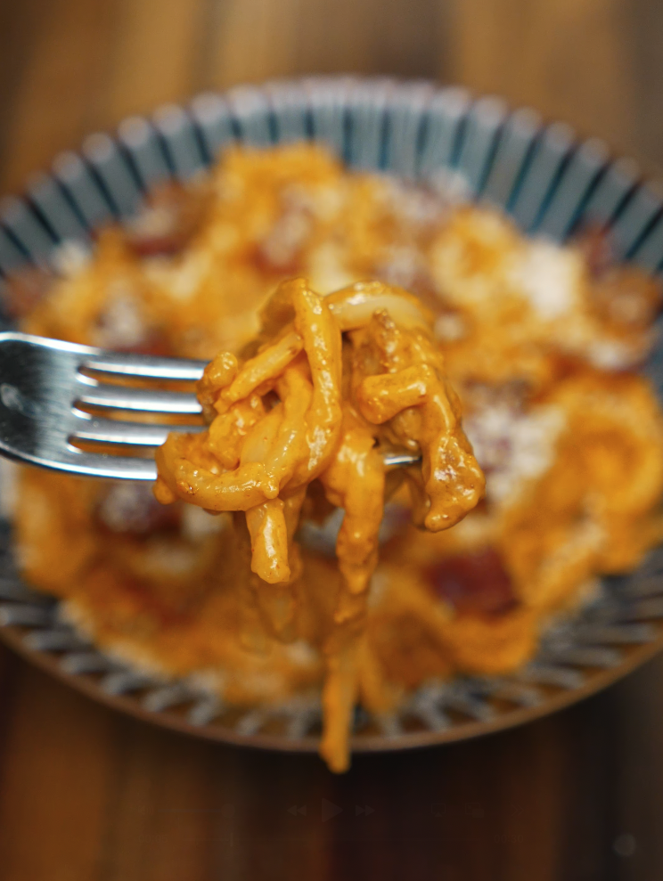 A close-up of a forkful of gochujang carbonara noodles lifted from a bowl, showing thick, glossy noodles coated in spicy sauce with crispy bacon pieces. The dish is creamy, bold, and rich in umami.