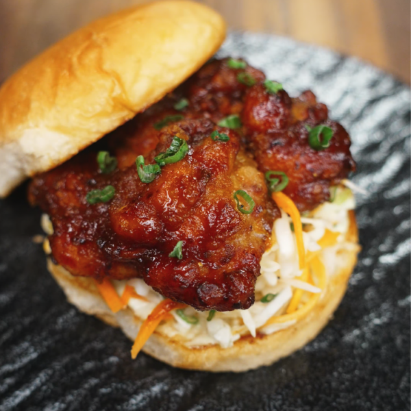 The assembled sandwich: a golden toasted bun loaded with crunchy cabbage slaw and a generous piece of glazed Korean fried chicken, topped with a sprinkle of green onions for freshness.