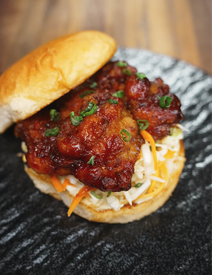 The assembled sandwich: a golden toasted bun loaded with crunchy cabbage slaw and a generous piece of glazed Korean fried chicken, topped with a sprinkle of green onions for freshness.