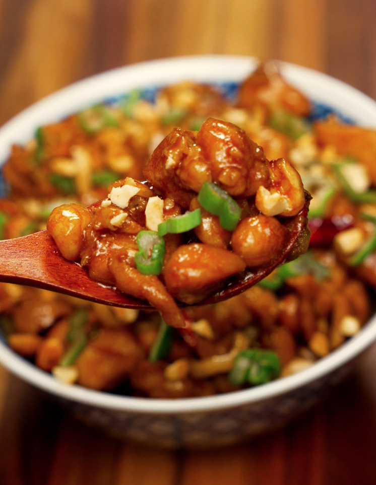 A wooden spoon lifts a saucy bite of Kung Pao Chicken Noodles with chunks of chicken, peanuts, and chopped scallions. The glossy sauce glistens under the light, showcasing texture and richness.