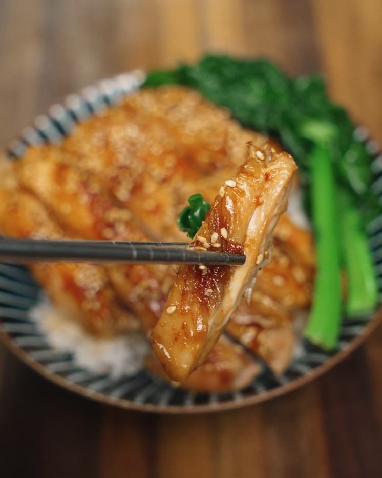 A juicy slice of honey garlic chicken lifted with chopsticks, showcasing the glossy glaze and sesame seed topping. Set against a backdrop of fluffy white rice and blanched green vegetables, this Asian-inspired chicken dish is perfect for meal prep, dinner bowls, or a healthy weeknight dinner. The chicken is tender, flavorful, and coated in a sticky-sweet sauce.