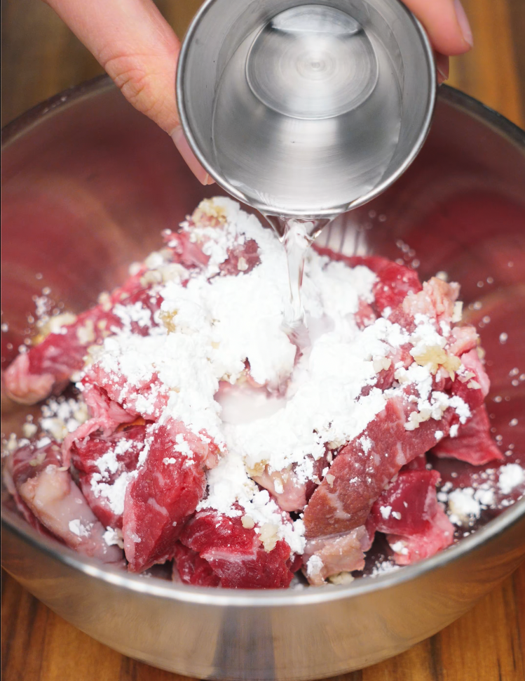 Thinly sliced beef in a metal bowl coated with cornstarch, garlic, and water, being prepared for deep-frying to achieve a crispy coating.