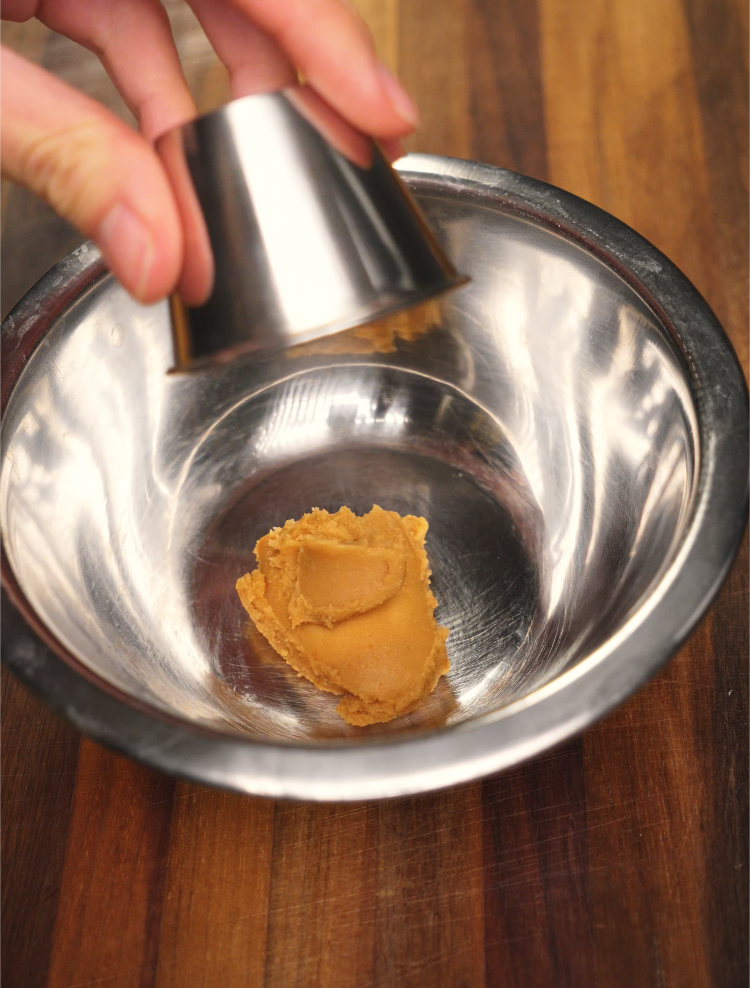 A spoonful of golden miso paste being added to a metal mixing bowl on a wooden surface, serving as the base ingredient for a savory glaze.
