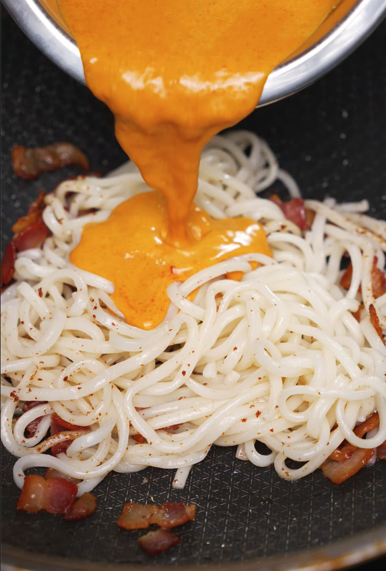 A rich, orange gochujang carbonara sauce being poured onto a bed of cooked udon noodles and crispy bacon. This Korean fusion pasta dish blends creamy, spicy, and savory elements into a bold, comforting meal.