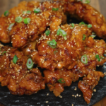 A heaping plate of honey butter chicken tenders stacked on a black ceramic dish, topped with scallions and sesame seeds. The golden glaze glistens on every piece, making this dish look both indulgent and irresistible—ideal for social media food content, fried chicken recipe blogs, or comfort food features.
