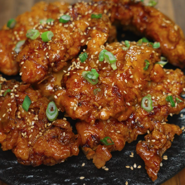 A heaping plate of honey butter chicken tenders stacked on a black ceramic dish, topped with scallions and sesame seeds. The golden glaze glistens on every piece, making this dish look both indulgent and irresistible—ideal for social media food content, fried chicken recipe blogs, or comfort food features.