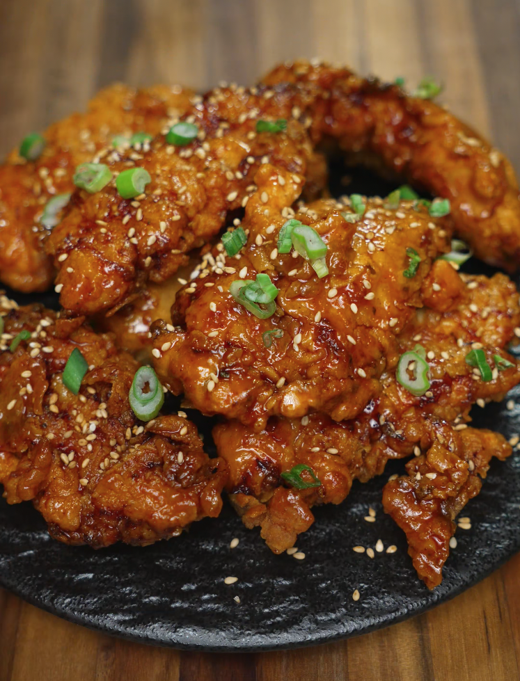 A heaping plate of honey butter chicken tenders stacked on a black ceramic dish, topped with scallions and sesame seeds. The golden glaze glistens on every piece, making this dish look both indulgent and irresistible—ideal for social media food content, fried chicken recipe blogs, or comfort food features.