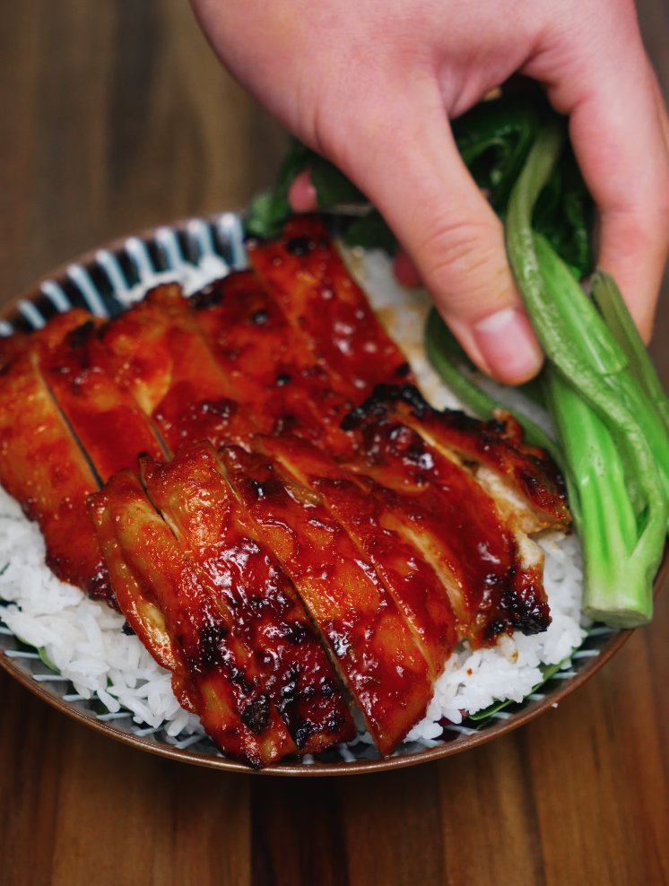 A hand placing bright green bok choy into a bowl of freshly cooked white rice topped with sliced BBQ gochujang chicken. The dish is almost ready to serve, showcasing meal prep ease and a healthy dinner combination of protein and greens with bold Korean flavor. A perfect fit for weeknight meals or Asian-inspired meal prep bowls.