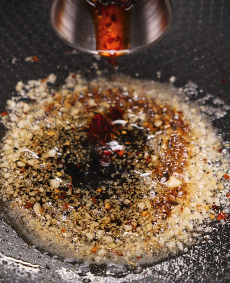 Hot chili oil being poured into a sizzling pan of minced garlic and melted butter — base for chili garlic butter noodles.